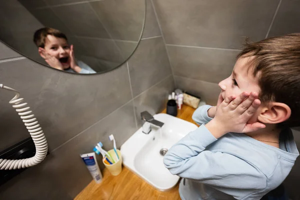 Boy washes with funny face holding cheeks in mirror at bathroom.