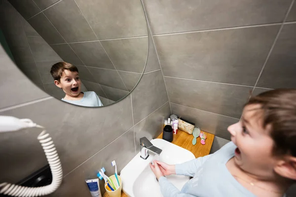 Boy washes with funny face in mirror at bathroom.