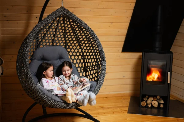 两个姐姐穿着睡衣 穿着针织袜子 靠着壁炉舒服地挂着椅子 在舒适的小木屋里看书 农村生活 — 图库照片