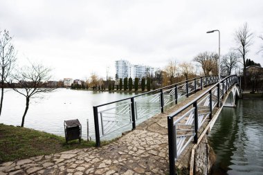Göl kenarındaki yaya köprüsünün arka planında cam bina çok katlı bir ev var..
