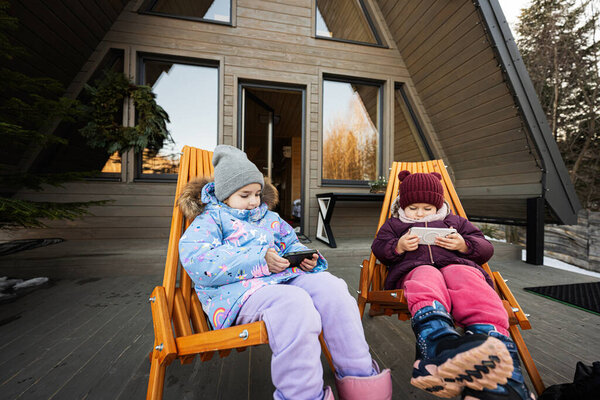 Two little girls sit on chairs at terrace off grid tiny house in the mountains and watching cartoons on mobile phones.