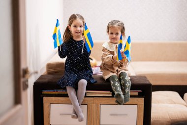 Two sisters are sitting on a couch at home with swedish flags on hands. Sweden children girls with flag .