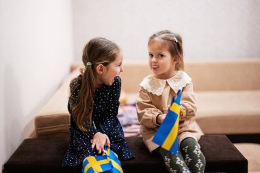Two sisters are sitting on a couch at home with swedish flags on hands. Sweden children girls with flag .