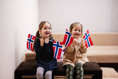 Two sisters are sitting on a couch at home with norwegian flags on hands. Norway children girls with flag .