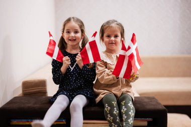 Two sisters are sitting on a couch at home with danish flags on hands. Denmark children girls with flag .