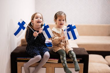 Two sisters are sitting on a couch at home with finnish flags on hands. Finland children girls with flag .