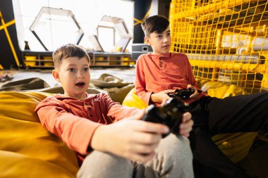 Oyun konsolu oynayan iki kardeş, oyun merkezinde sarı puf üzerinde oturuyorlar..