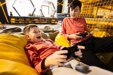 Oyun konsolu oynayan iki kardeş, oyun merkezinde sarı puf üzerinde oturuyorlar..