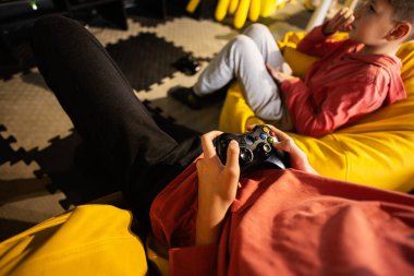 Oyun konsolu oynayan iki kardeş, oyun merkezinde sarı puf üzerinde oturuyorlar..