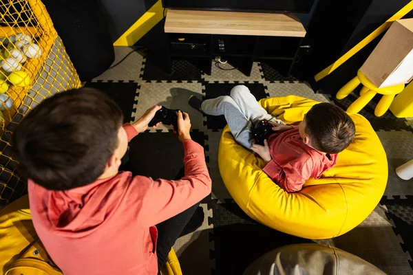 stock image Two brothers playing video game console, sitting on yellow pouf in kids play center.