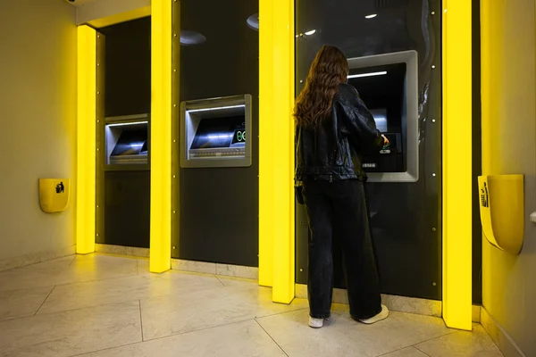在黄色自动取款机上从信用卡取款的年轻黑发姑娘身后 免版税图库图片