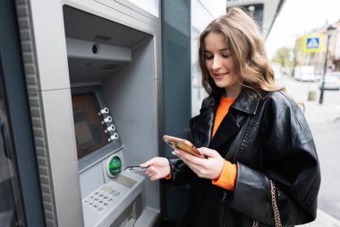 Deri ceketli genç bir kadın akıllı telefonlara bakarken ATM 'ye kredi kartı sokuyor..