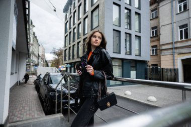 Deri ceketli genç kız şehir merkezinde poz vermiş, elinde el çantası ve telefonuyla kredi kartı var..