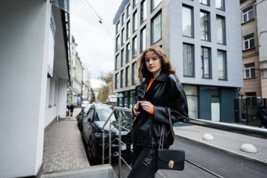 Deri ceketli genç kız şehir merkezinde poz vermiş, elinde el çantası ve telefonuyla kredi kartı var..