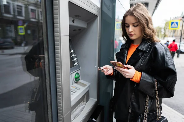 身穿皮夹克的年轻女子一边在室外自动取款机上插入信用卡 一边看智能手机 免版税图库照片