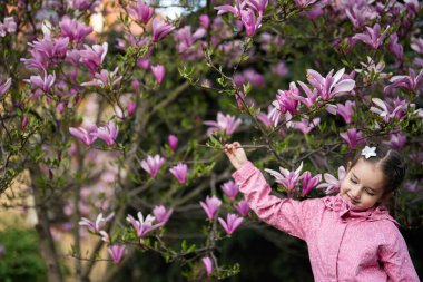 Pembe ceketli anaokulu kızı manolya çiçekli ağacın yanında güzel bir bahar gününün tadını çıkarıyor. İlkbahar aktiviteleri.