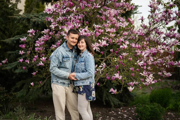 恋爱中的温情夫妻穿着牛仔裤夹克 在木兰花盛开的树旁享受着美丽的春日 — 图库照片