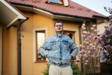 Gözlüklü ve kot ceketli şık bir adamın portresi evine karşı poz vermiş..