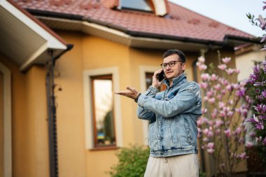 Gözlüklü ve kot ceketli şık bir adamın portresi, evine cep telefonu dayamış ve sol tarafta el hareketi yapıyor..