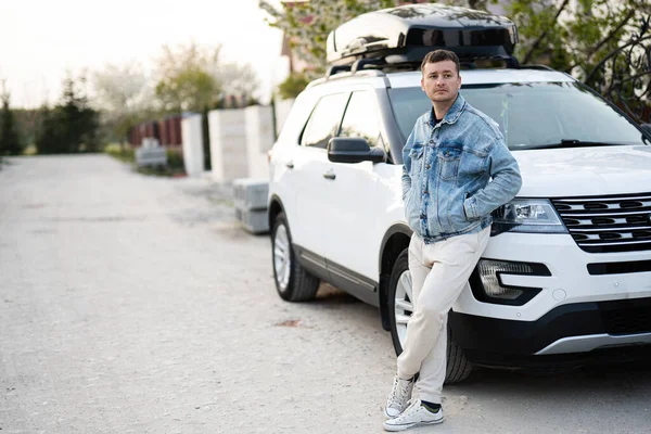 Man in jeans jacket standing near white american suv car with roof rack box.