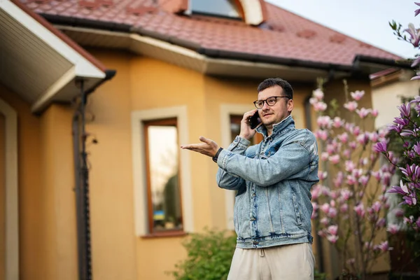 Gözlüklü ve kot ceketli şık bir adamın portresi, evine cep telefonu dayamış ve sol tarafta el hareketi yapıyor..