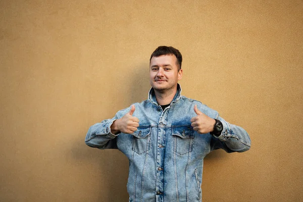 Portrait of stylish man in jeans jacket against yellow wall show thumbs up.