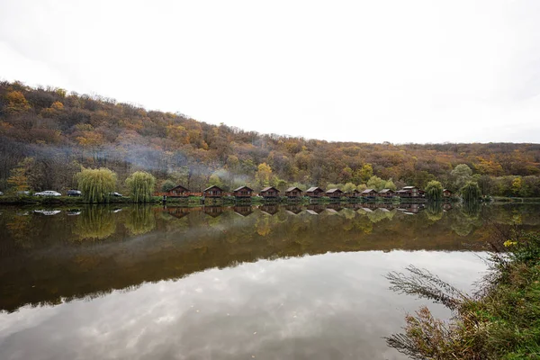 Country landscape. Wooden houses with smoke on the lake. Reflections of a building in the water on a autumn day
