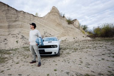 Şapkalı yakışıklı adam kariyerinde beyaz kaslı arabasının yanında duruyor..