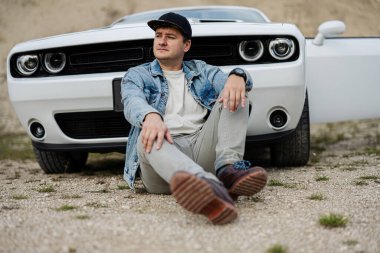 Kariyeri boyunca beyaz kaslı arabasının yanında oturan kot ceketli ve şapkalı yakışıklı bir adam..