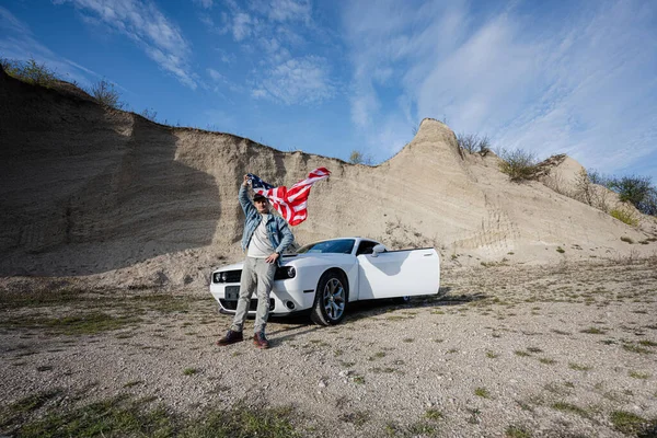 ジーンズジャケットとキャップでハンサムな男でアメリカ国旗近く彼の白いアメリカの筋肉車でキャリア — ストック写真