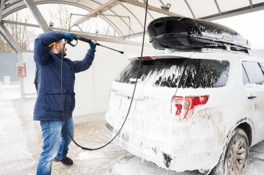 Man washing high pressure water american SUV car with roof rack at self service wash in cold weather.