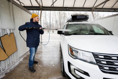 Man washing high pressure water american SUV car with roof rack at self service wash in cold weather.