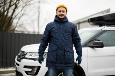 Man driver wear jacket and yellow hat against his american SUV car with roof rack in cold weather.