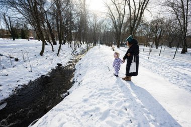 Mother and child on a sunny frosty winter day in the park feed ducks on frozen river.