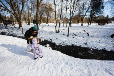 Mother and child on a sunny frosty winter day in the park feed ducks on frozen river.