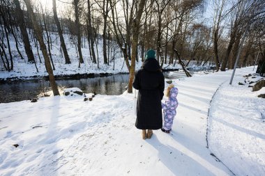 Mother and child walking on a sunny frosty winter day in the park near river with ducks and birds.