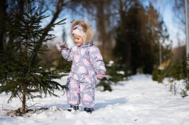 Baby girl wear child snowsuit on a sunny frosty winter day near Christmas tree.