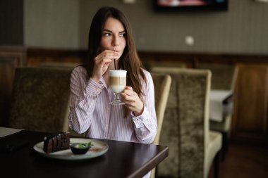 Genç, güzel esmer kadın, kafeteryada oturuyor, kapalı bir kafede ve pipetten latte içiyor.. 