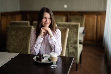 Genç, güzel esmer kadın kafeteryada oturuyor, kapalı bir kafede ve yüzünü buruşturuyor..