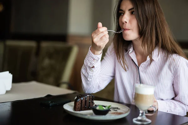 stock image Young beautiful brunette woman sit in coffee shop cafe restaurant indoors and eat chocolate brownie dessert cake, fork near the mouth.