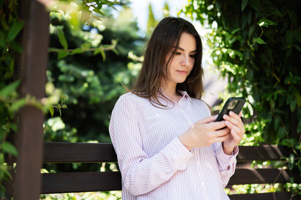 Young freelancer woman in shirt sit on bench in summer park outdoors rest and use mobile cell phone. 