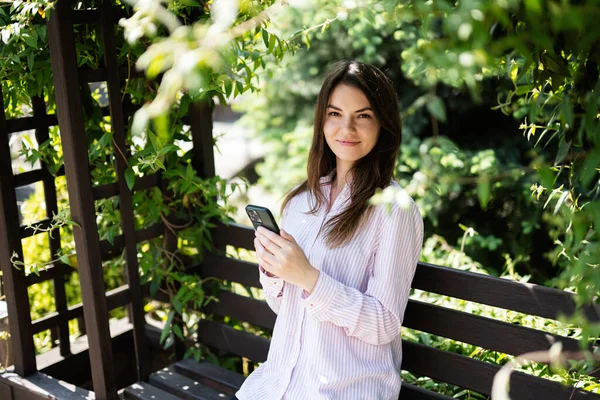 stock image Young freelancer woman in shirt sit on bench in summer park outdoors rest and use mobile cell phone. 