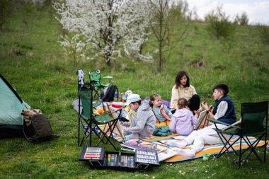 Mutlu genç aile, anne ve çocuklar bahçedeki bahar parkında piknik battaniyesinde eğleniyor ve eğleniyorlar..