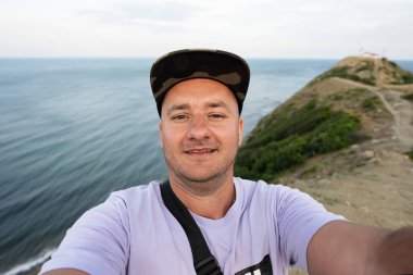 Kapüşonlu gülümseyen bir adamın portresi denizin arka planında selfie çekiyor. Cape Emine, Karadeniz kıyısı, Bulgaristan.