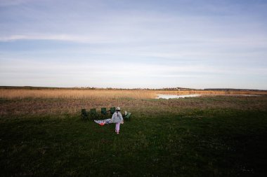 Hazırlanmış bir piknik alanına yaklaşan bir çocuğu görüntülemek. Geniş, açık bir alan ve berrak bir gökyüzünün altında sakin bir gölet tarafından çerçevelenmiş..