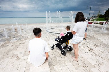 Bebek arabasında bir çocukla deniz kenarında kaliteli bir zaman geçirmek. Çeşmeler ve bulutlu bir gökyüzü huzurlu manzarayı çerçeveler..