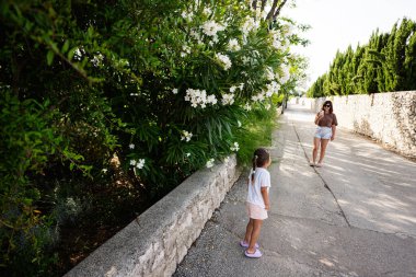 Hırvatistan 'ın Nin kentinde güneşli bir günde, huzurlu bir yürüyüş yolu boyunca çiçek açan kızının fotoğrafını çeken bir anne.