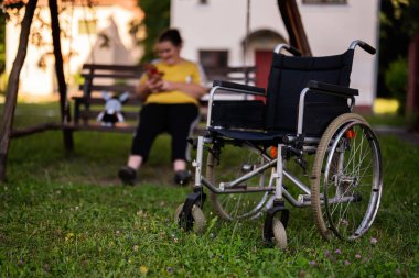 Huzurlu bir park ortamında ön planda boş bir tekerlekli sandalyeyle bankta oturan engelli bir kadın.