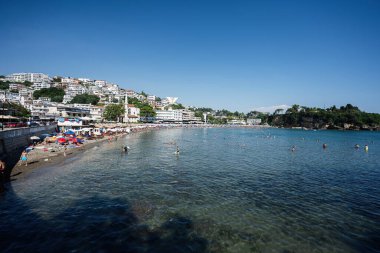 Ulcinj, Karadağ - 13 Temmuz 2024: Açık denizler, canlı şemsiyeler ve açık mavi gökyüzü altında kıyı kasabası bulunan kumlu bir plajda insanları yakalayan hareketli bir sahnedir.