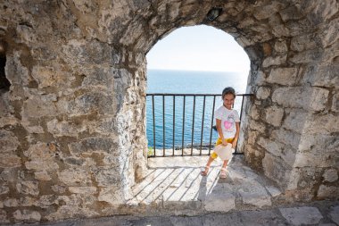 Genç bir kız açık mavi denize bakan taştan bir kemer yolunda duruyor, Karadağ 'ın Ulcinj kentinde güneşli bir kıyı manzarasının tadını çıkarıyor.